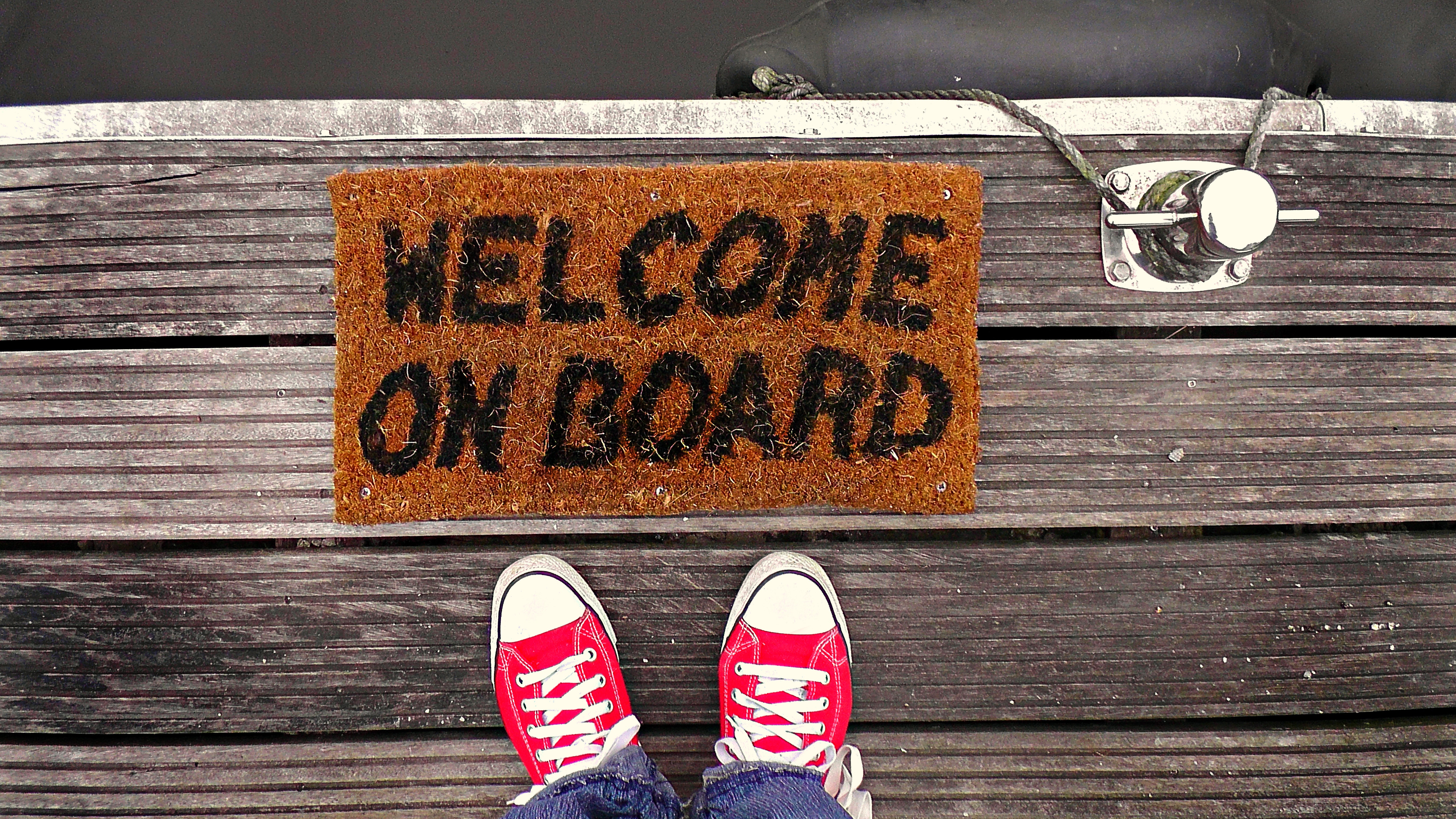 Red sneakers on a wooden floor that looks like a part of a boat in front of a doormat that says "WELCOME ON BOARD"